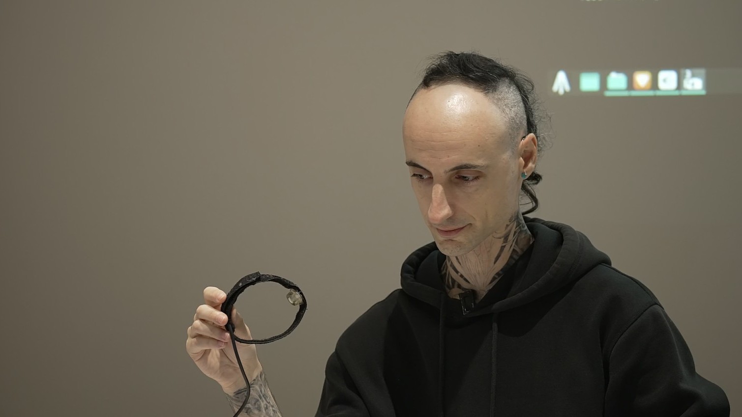 실내 워크숍 환경에서 검은 후드티를 입고 있는 사람이 테이블에 앉아 작은 원형 전자 장치를 손에 들어 자세히 살펴보고 있는 모습. 배경에는 흐릿한 화면과 아이콘이 보이며, 기술 시연이나 제작 과정을 설명하는 분위기다.